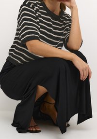 Gestreifter Strickpullover in Schwarz und Creme, kombiniert mit einem schwarzen, fließenden Rock mit einem seitlichen Schlitz, abgerundet durch schwarze Riemensandalen.