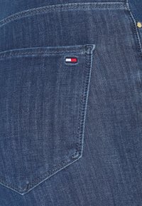 Vaqueros de mezclilla azul oscuro con un bolsillo trasero que presenta un pequeño logo en rojo, blanco y azul. Observa las costuras visibles y los detalles de la textura.