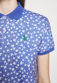 Polo bleu avec imprimé floral blanc et un petit logo de cheval vert brodé sur la poitrine, porté par une personne.