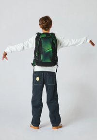 Sac à dos noir et vert avec un motif géométrique, sangles ajustables et plusieurs compartiments. Porté par une personne en tenue décontractée.