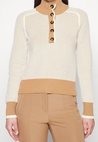 Kobieta wearing beżowy i kremowy sweter robiony na drutach z czterema guzikami z przodu, zestawiony z spodniami w kolorze kamelowym.