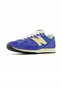 Sininen ja violetti urheilutossu, jossa on teksturoitu päällinen, kreemiväriset nauhat ja keltaiset yksityiskohdat. Varustettu mustalla kumipohjalla ja klassisella mallilla.