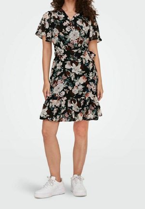 Femme portant une robe portefeuille noire à fleurs avec ourlet volanté et baskets blanches, se tenant devant un fond blanc uni.