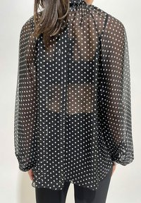 Donna che indossa una blusa nera trasparente con pois bianchi, che mostra uno strato sottostante nero, in piedi davanti a uno sfondo semplice e chiaro.