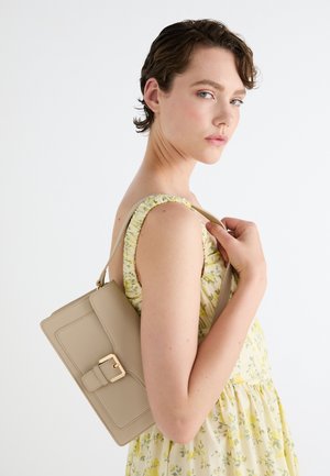 Junge Frau in einem gelben geblümten Kleid hält eine beige rechteckige Handtasche über der Schulter vor einem einfarbigen weißen Hintergrund.
