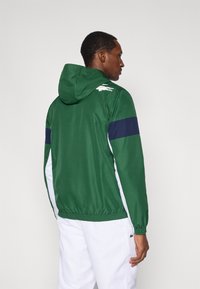 Zielona kurtka z kapturem z granatowym paskiem na ramionach, elastycznym dołem, białymi panelami bocznymi oraz logo na plecach. Wykonana z lekkiego materiału.
