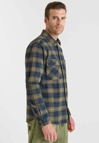 Man in een blauw en olijfgroen geruite knoopshirt met borstzakken, zittend op een zijlijn tegen een effen witte achtergrond.