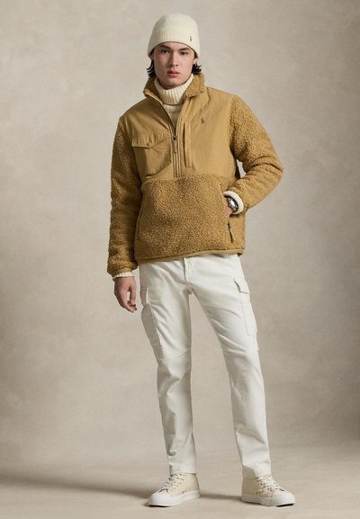 Μπεζ φλις μπουφάν με φερμουάρ μπροστά και δύο τσέπες στο στήθος, συνδυασμένο με λευκό παντελόνι cargo και εκρού σκουφάκι. Casual, με υφή εμφάνιση.