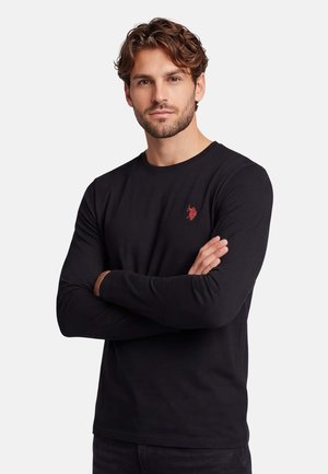 Mann mit welligem Haar, der ein schwarzes Langarmshirt mit einem kleinen roten bestickten Logo auf der Brust trägt, die Arme verschränkt.