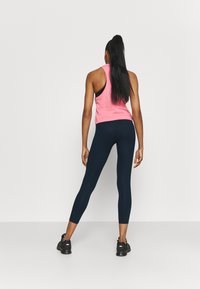 Kvinna sedd bakifrån stående med benen isär, iförd en rosa ärmlös topp, svarta leggings och svarta sportskor mot en neutral bakgrund.