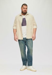 Beige corduroy shirt over een wit T-shirt met een paarse vierkante grafiek, gecombineerd met blauwe jeans en witte sneakers op een grijze achtergrond.
