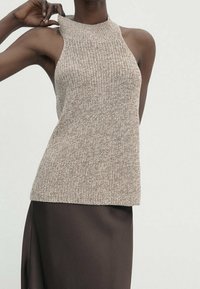 Beige ärmelloses Strickoberteil mit hohem Halsauschnitt. Gewebte Textur mit lässiger Passform. Kombiniert mit einem glatten dunkelbraunen Rock.