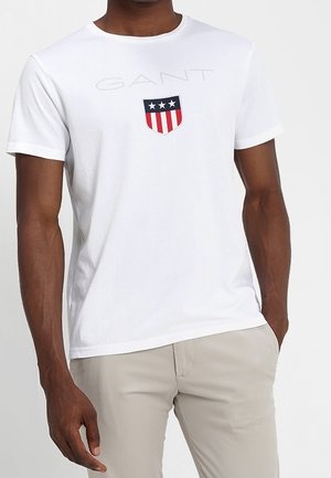 Mężczyzna ubrany w białą koszulkę GANT z logo tarczy zawierającym trzy gwiazdy i czerwone pionowe paski, w połączeniu z jasnobeżowymi spodniami.
