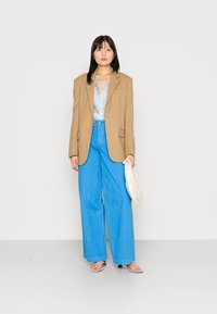 Blazer bege oversized sobre um top floral azul claro, combinado com calças largas de cintura alta em azul brilhante e sandálias simples. Segurando uma bolsa branca.