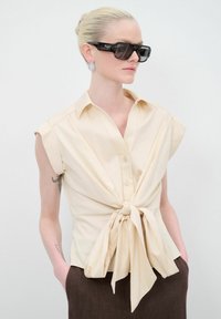 Blusa beige a maniche corte con un fronte annodato, colletto e bottoni. Il modello l'ha abbinata a un pantalone scuro a vita alta e occhiali da sole neri oversize.