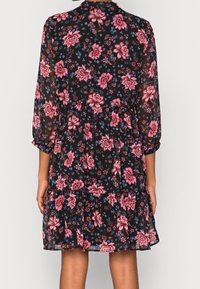 Femme portant une robe noire avec des motifs floraux roses et bleus, à manches longues et avec un ourlet en volants, vue de dos.