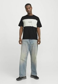 Camiseta de color bloque negro y verde pálido con el texto "Original Studio", acompañada de jeans de mezclilla azul claro y zapatillas deportivas negras.