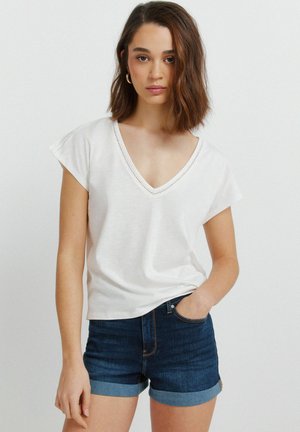 Jonge vrouw met schouderlang bruin haar, gekleed in een wit T-shirt met V-hals en donkerblauwe opgerolde denim shorts, poserend met één hand in haar zak.