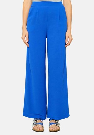 Frau trägt hochtaillierte, weit geschnittene blaue Hosen mit Taschen und silbernen Riemchensandalen, steht vor einem schlichten weißen Hintergrund.