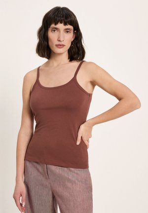 Woman with dark hair wearing a brown sleeveless top and light mauve pants, standing with one hand on her hip against a plain background.