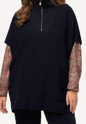 Kobieta nosząca czarny, prążkowany, dzianinowy ponczo z przednimi kieszeniami i kołnierzem na pół suwaka, na długim rękawie koszuli w wzór paisley.