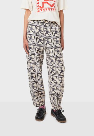 Pantaloni leggeri a fantasia in beige con disegni geometrici scuri, vestibilità ampia, vita elastica, abbinati a scarpe nere.