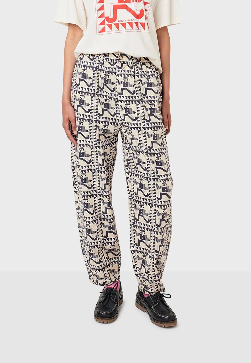 Persoon in crème broek met zwart geometrisch dierenprint, wit t-shirt met rode grafiek, zwarte schoenen en roze sokken, staand op een grijze vloer.