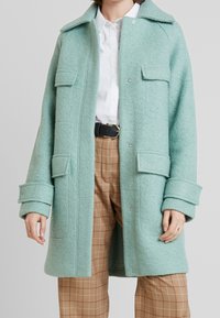 Cappotto in lana verde menta con colletto classico, due tasche frontali e chiusure a pressione, abbinato a pantaloni a quadri beige e una camicia bianca.