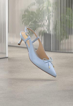 Zapato de tacón bajo tipo kitten azul claro con tira trasera, punta puntiaguda y detalle de lazo, flotando sobre un suelo gris, con una cerca metálica difuminada y plantas de fondo.