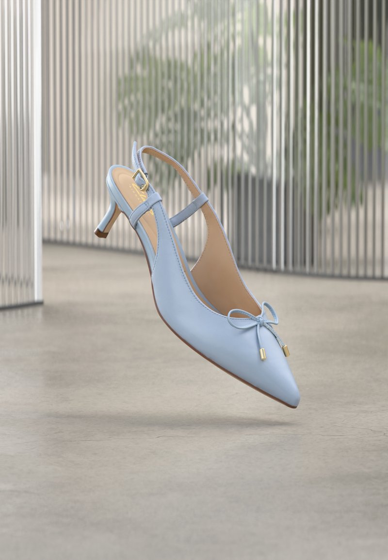 Világoskék pántos cipő kis sarokkal, vékony hegyes orral és masni díszítéssel, szürke padló felett lebegve, háttérben elmosódott fém kerítés és növények.