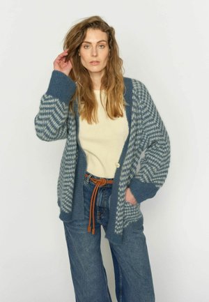 Vrouw met lang golvend haar, gekleed in een blauwe gebreide cardigan met patroon, een crèmekleurige trui, high-waisted jeans en een bruine knoopriem, staand tegen een witte achtergrond.