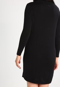 Abito nero lavorato a maglia con maniche lunghe, scollo alto e vestibilità rilassata, caratterizzato da un orlo fino al ginocchio e una texture liscia e opaca.