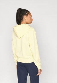 Κίτρινο φούτερ με κουκούλα και άνετη εφαρμογή, με ριμπ τελείωμα και μανσέτες. Φορεμένο με μπλε σκούρο παντελόνι, εμφανίζοντας ένα πλεκτό χτένισμα από την πίσω πλευρά.