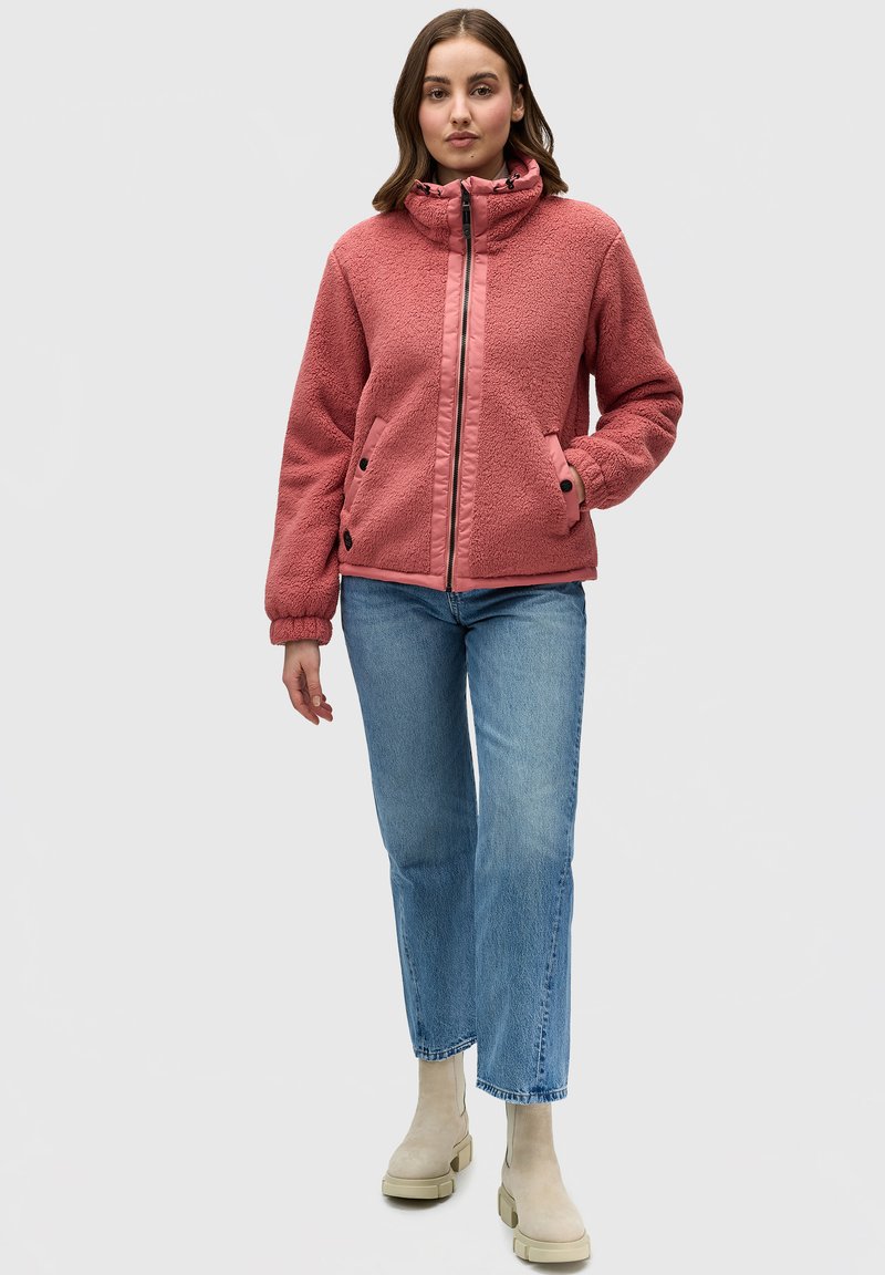 Giacca in pile di un delicato rosa con colletto alto, chiusura con zip, polsini elasticizzati e due tasche laterali; abbinata a jeans dritti blu chiaro e stivali beige.