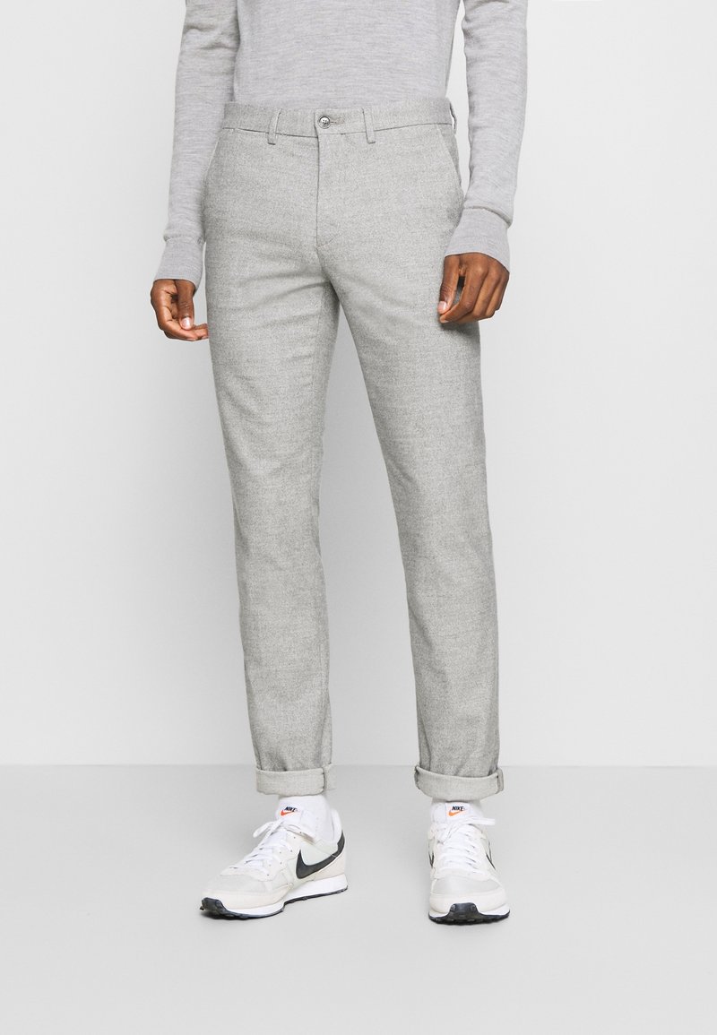 Person klädd i ljusgrå slim-fit byxor med uppvikt mudd, tillsammans med vita Nike-sneakers och en ljusgrå långärmad topp.