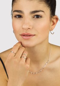Woman with dark hair in a bun wearing silver hoop earrings, a delicate necklace, and three silver rings, touching her chin thoughtfully.