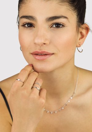 Frau mit dunklem Haar im Dutt, die silberne Creolen, eine zarte Halskette und drei silberne Ringe trägt, und nachdenklich ihr Kinn berührt.