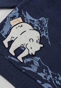 Tessuto di cotone blu scuro con un ricamo bianco che rappresenta un orso polare e grafiche di montagne blu chiaro. Include un piccolo etichetta con il marchio.