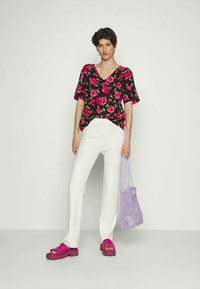 Pessoa em pé contra uma parede simples, vestindo uma blusa preta floral, calças brancas, sandálias de plataforma rosas e segurando um saco de malha roxo.
