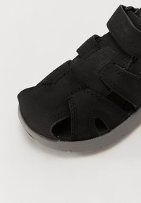 Sandalia negra con parte superior de ante, múltiples aberturas y correa ajustable. Presenta una suela de goma gris para tracción y comodidad.