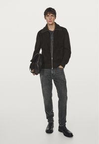 Młody mężczyzna w czarnej zapinanej na zamek bluzie, szarych dżinsach i czarnych botach, trzymający złożoną czarną torbę na tle białym.