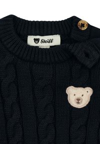 Jersey de punto azul marino con patrón de trenzas, que presenta un parche de oso y un botón beige en el hombro. La etiqueta muestra "Steiff".
