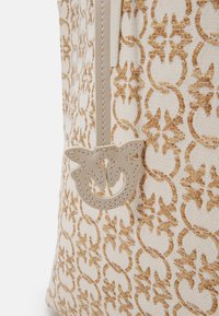 Sac en tissu beige et crème avec un motif floral et circulaire tissé marron et un patch logo en cuir beige cousu sur le côté.