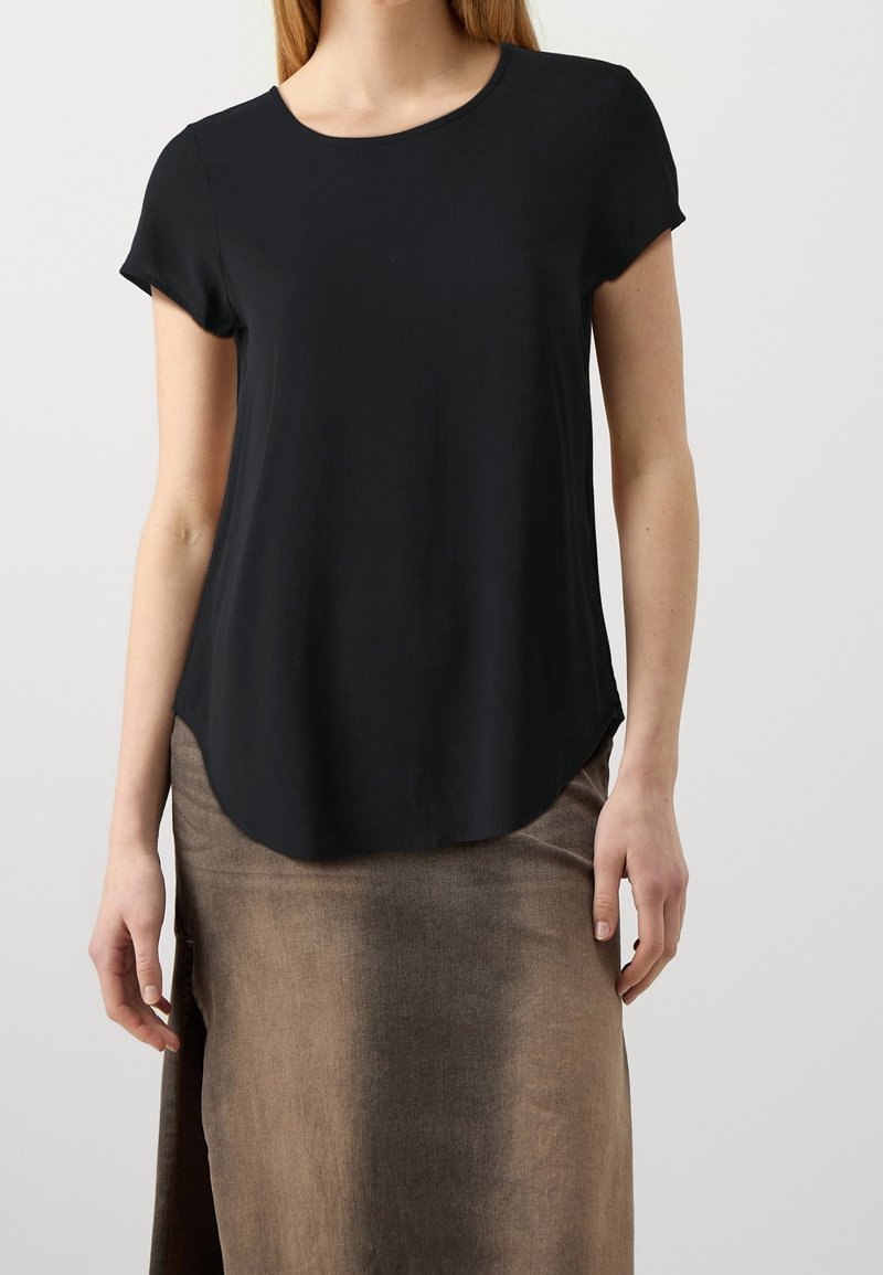 Femme portant un haut noir uni à manches courtes et une jupe marron avec une fente latérale, debout devant un fond clair.