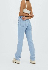 Femme de dos face à la caméra, portant un jean bleu clair, un crop top blanc et des baskets beiges, le tout devant un fond blanc uni.