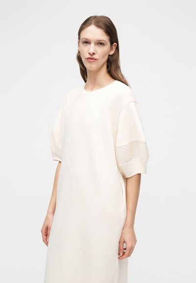 Νεαρή γυναίκα με ανοιχτό καστανά μαλλιά φοράει ένα αέρινο, εκρού φόρεμα με μανίκια μέχρι τον αγκώνα και φουσκωτά, στέκεται μπροστά σε απλό φωτεινό φόντο.