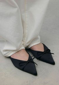 Ballerines en satin noir à bout pointu avec un petit nœud sur le cou-de-pied, portées avec un pantalon blanc à revers. Texture lisse, design épuré.