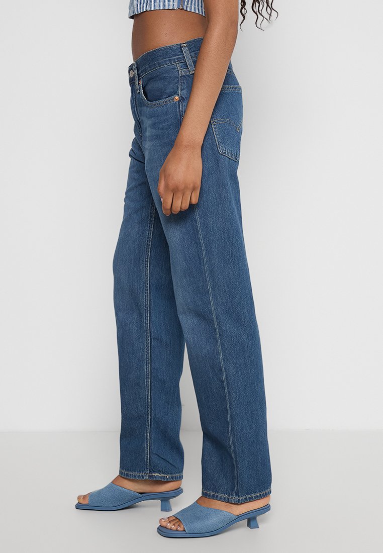 Jeans en denim bleu à jambes larges avec une taille haute, design classique à cinq poches et détails de couture minimaux. Associés à des talons ouverts bleus.