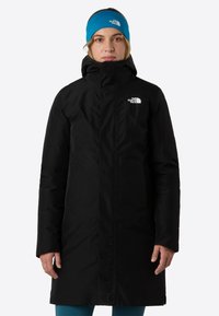 Čierna zateplená parka s vysokým golierom, predn�ým zipsom a bočnými vreckami. Nosená s modrým čelenkou. Hladká textura a minimalistický dizajn.