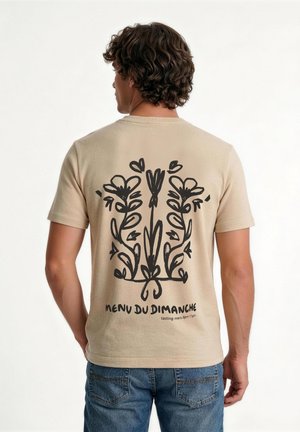 Homme aux cheveux bouclés portant un t-shirt beige avec des motifs floraux noirs et le texte « Menu du Dimanche » au dos, associé à un jean bleu.